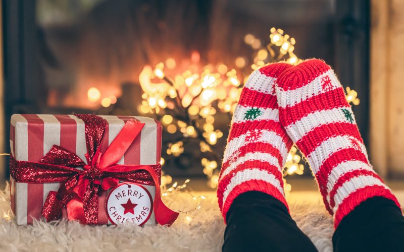 Feet in Christmas socks and gift box in front of the fireplace, close-up, relaxing at home, cozy background, copy space.